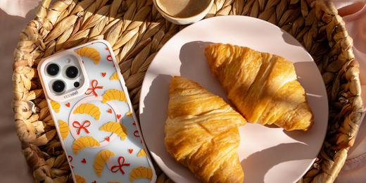  EcoBlvd eco-friendly phone case with croissant and bow patterns, placed beside real croissants and coffee on a woven tray.