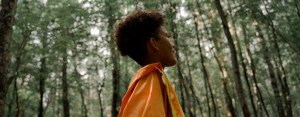 A person wearing an orange windbreaker looking up at the trees while in a lush forest of tall trees.